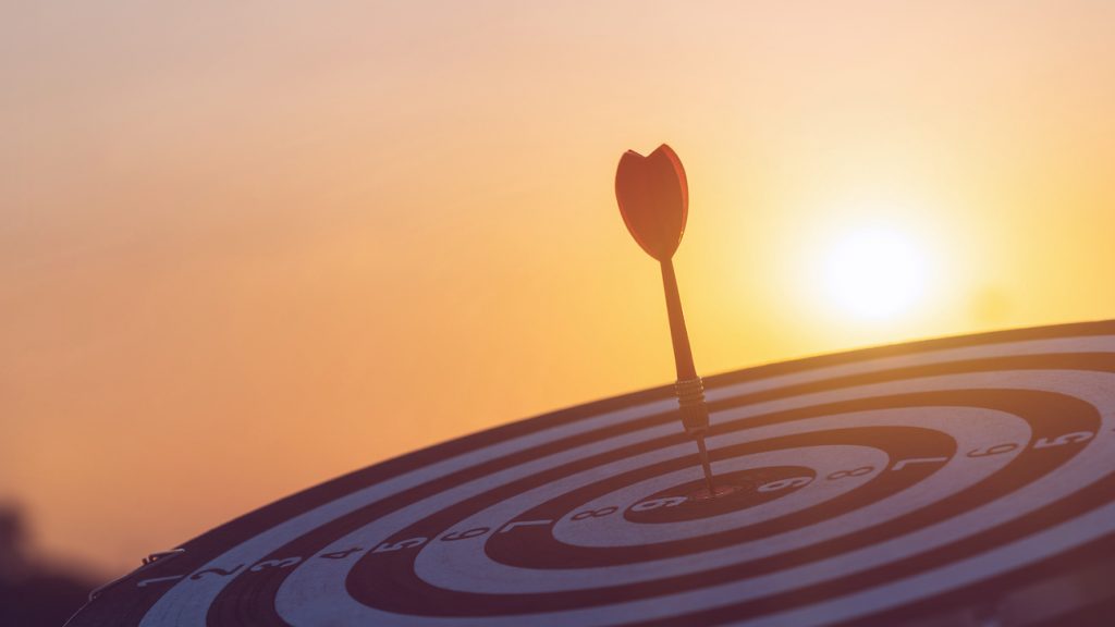 Close up red dart arrow hitting target center dartboard on sunset background. Business targeting and focus concept