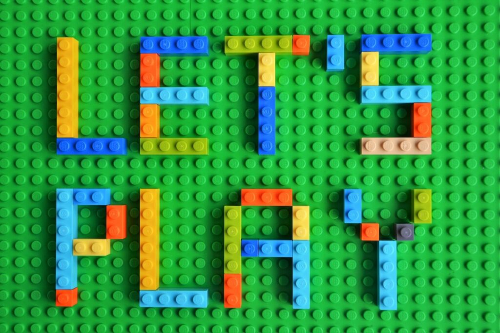 LET'S PLAY bunte Aufschrift auf dem grünen Sockel/Platte/Brett. Die Buchstaben sind aus Legosteinen gefertigt.